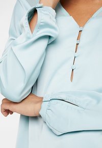 Blouse bleu clair avec un décolleté en V, trois boutons décoratifs et des manches froncées avec une texture subtile.