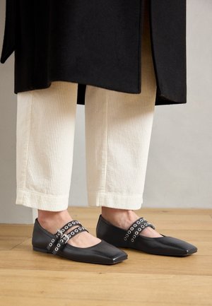 Osoba ubrana w kremowe spodnie sztruksowe i czarne płaskie buty z podwójnymi paskami, stojąca na drewnianej podłodze.