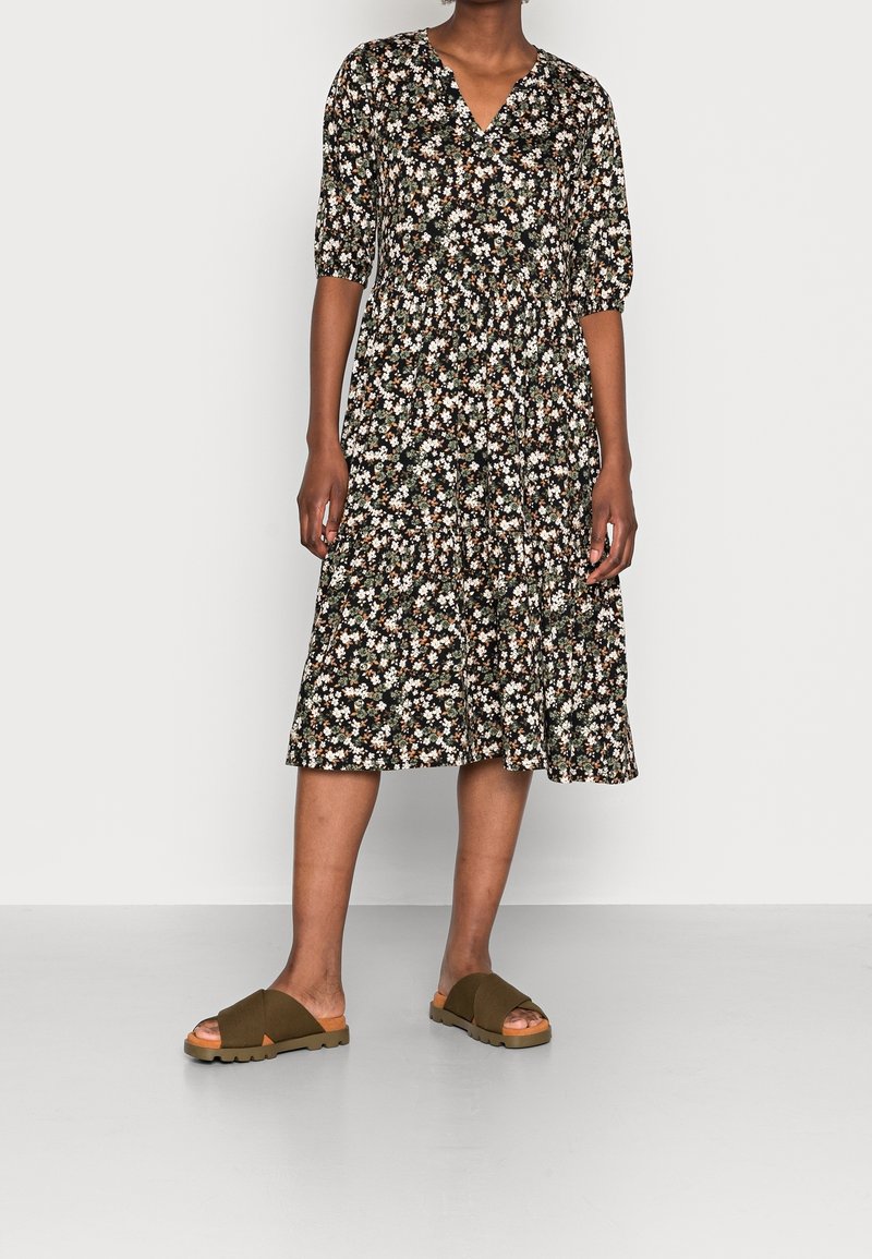 Robe noire à fleurs avec un col en V, des manches courtes bouffantes et une jupe à volants. Assortie à des sandales marron à brides croisées sur un fond clair.