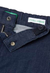 Joggers de mezclilla azul oscuro con cierre de cremallera, botón y cintura elástica, con detalles en etiqueta verde y herrajes plateados.