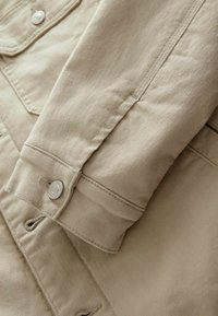 Beige bomuldsjakke med knaplukning, der har et tekstureret stof, broderede sømme og en detaljeret manchet med knap.