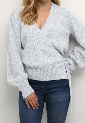 Femme portant un pull cache-cœur gris clair avec des poignets et un ourlet côtelés, noué sur le côté, associé à un jean bleu foncé, visible du cou jusqu'aux hanches.
