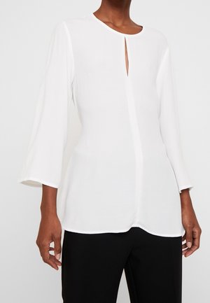 Femme portant un chemisier blanc à manches longues avec une petite fente devant et un pantalon noir, debout devant un fond uni.