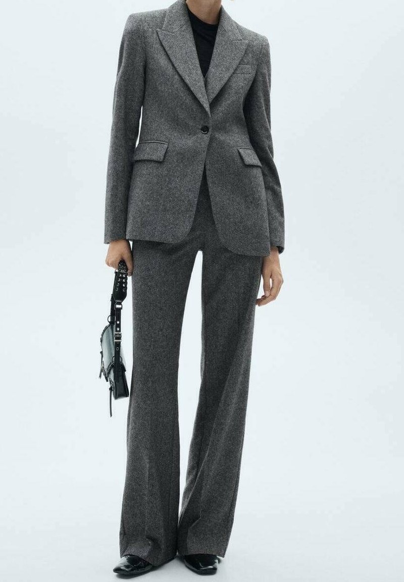 Personne portant un blazer gris ajusté avec un pantalon large assorti, des chaussures noires et tenant un petit sac à main noir.