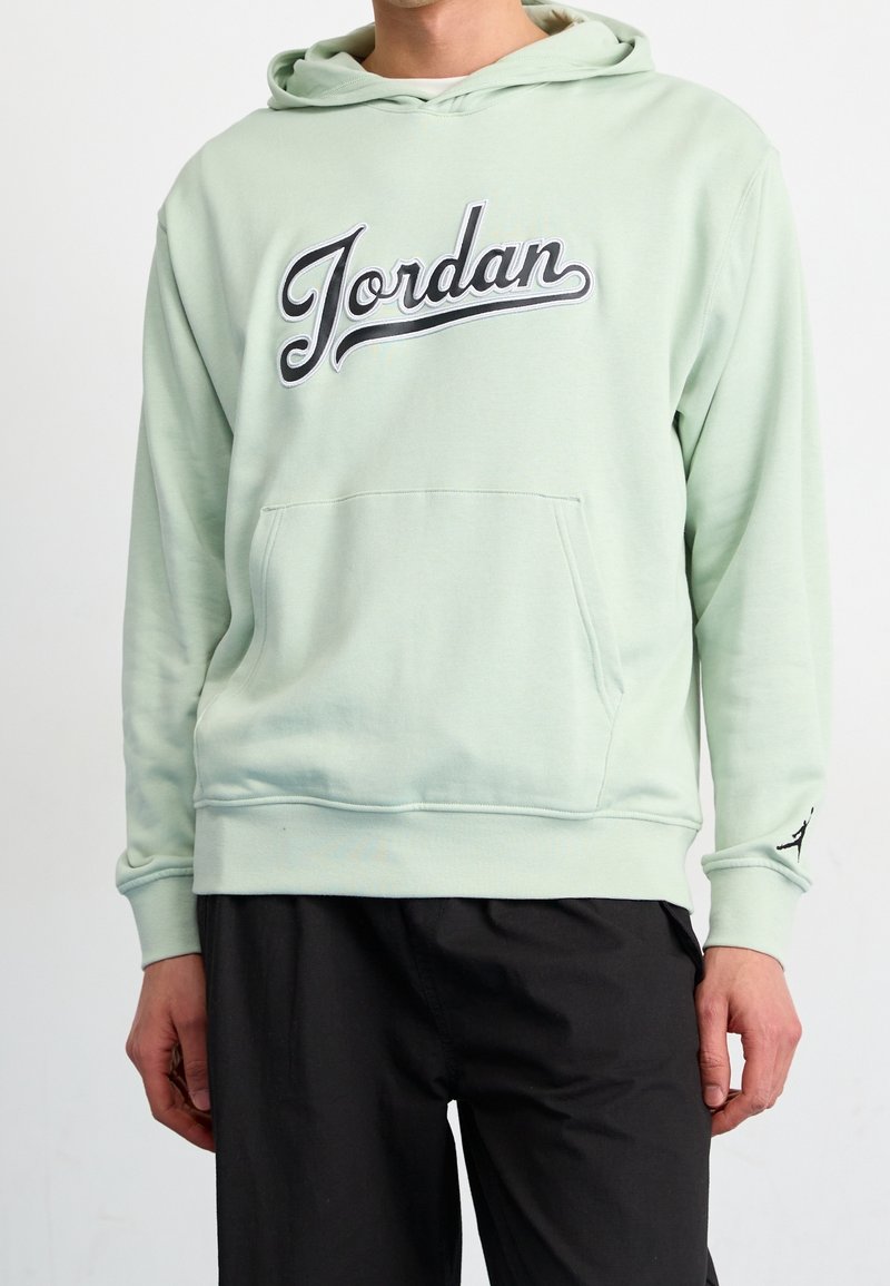 Personne portant un sweat à capuche vert clair avec le texte « Jordan » sur la poitrine et un pantalon noir.