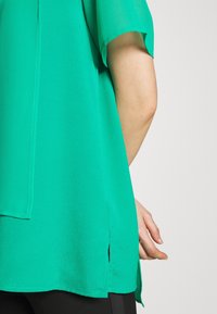 Blusa verde a maniche corte con una texture liscia, fessure laterali e una vestibilità comoda, indossata su pantaloni neri, mostrando parte di una mano.