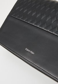 Fekete bőr kézitáska, amelynek a felső fele steppelt mintázattal, az alsó része pedig sima felülettel rendelkezik, ezüst 'Calvin Klein' logóval.