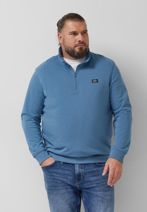 Mann mit Bart und zurückgebundenem Haar, der einen blauen Sweatshirt mit Reißverschluss und Jeans trägt, steht mit einer Hand in der Tasche vor einem schlichten Hintergrund.