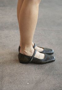 Zwarte, vierkante balletflats met een gestructureerd slangenhuidpatroon en een dunne band over de wreef, tegen een neutrale vloer.