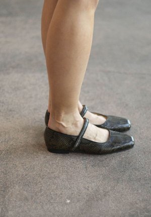 Ballerine nere con punta quadrata, in pelle martellata a forma di serpente e una sottile fascia sulla parte superiore del piede, posizionate su un pavimento neutro.