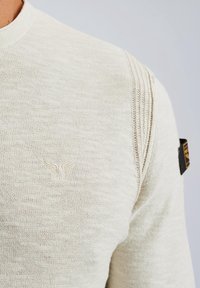 Pull en maille beige clair avec un col rond, présentant un logo brodé discret sur la poitrine et des détails côtelés texturés sur les épaules.