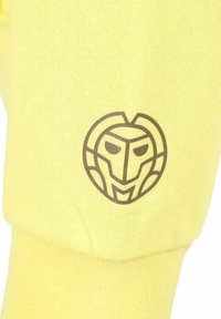 Gele stof met een gestructureerd oppervlak, voorzien van een bruin logo dat lijkt op een gestileerd masker of gezicht, geplaatst op een mouw.