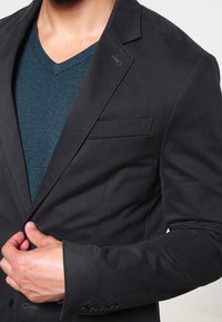 Mężczyzna dostosowujący guziki w czarnym żakiecie noszonym na turkusowym swetrze w serek, pokazujący górną część torsu i zbliżenie na rękę.