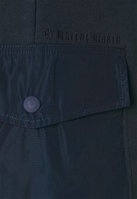 Mörk marinblå tyg med en texturerad övre del och en slät nedre del. Innehåller en lockficka säkrad med en blå knapp och präglad logotyp.