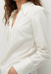 Chemise blanche boutonnée en tissu lisse, avec col pointu, manches longues et une poche poitrine unique.