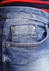 Ljusblå denimjeans med en liten bakficka med silvernitar och kontrasterande bruna sömmar. Tyget har en blekt textur.