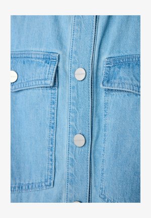 Camisa de mezclilla azul claro con dos bolsillos frontales, botones a presión plateados y costuras visibles. La tela muestra una textura sutil.