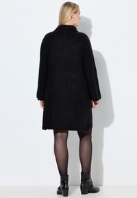 Zwarte geribbelde lange cardigan met een brede kraag en zijsplitten, gemaakt van een getextureerde gebreide stof. Draagt over zwarte maillots en enkellaarzen.