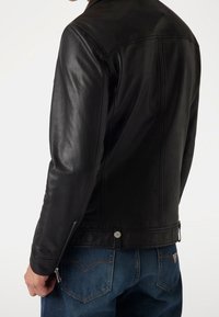 Mann in einer taillierten schwarzen Lederjacke mit Reißverschlusstaschen und blauen Jeans, von hinten vor einem weißen Hintergrund gezeigt.