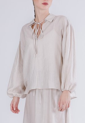 Mujer vistiendo una blusa arrugada beige claro con mangas largas abullonadas y detalle de lazo en el cuello, combinada con pantalones holgados a juego.