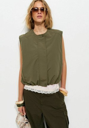 Vrouw draagt mouwloos olijfgroen gewatteerd vest, wit topje met kanten rand, olijfgroene cargobroek, zonnebril, houdt een handtas van slangenleer met armbanden om de polsen vast.