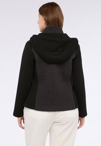 Vrouw gezien van achteren, gekleed in een zwarte jacket met een textuur aan de achterkant en een capuchon, gecombineerd met witte broek tegen een plain achtergrond.