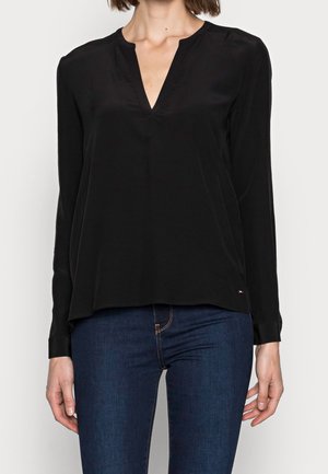 Kvinde iført en sort bluse med lange ærmer og V-hals samt mørkeblå jeans, stående foran en ensfarvet lys baggrund.