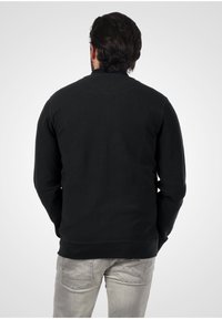 Schwarzer Sweatshirt mit rundem Ausschnitt und gerippten Bündchen, kombiniert mit hellgrauen Jeans, die auf den hinteren Taschen einen Zickzackstich aufweisen.