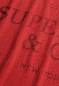 Czerwona tkanina o gładkiej fakturze z ciemnymi nadrukowanymi literami, w tym "SUPER" i "& CO", z napisem "NEW YORK" na dole.