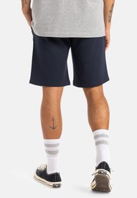 Pantalones cortos deportivos de color azul marino, mezcla de algodón, con cinturilla acanalada y textura suave. Calcetas blancas hasta la rodilla con rayas grises y zapatillas negras.