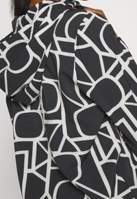 Schwarzer Stoff mit einem markanten, abstrakten weißen geometrischen Muster. Verfügt über eine Kapuze und eine fließende Silhouette, die verschiedene Formen und Linien präsentiert.