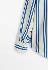 Camicia a maniche lunghe crema con strisce verticali blu e polsini abbottonati su uno sfondo bianco.