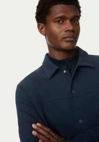 Giacca camicia in tessuto blu navy con chiusura a bottoni, colletto e superficie liscia. Presenta un design pulito e dettagli sottotono.