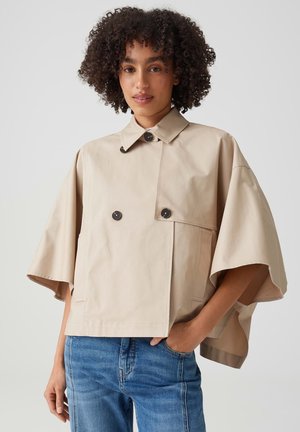 Giovane donna con capelli ricci che indossa un trench beige a maniche corte e jeans blu, in piedi con una mano in tasca su uno sfondo semplice.