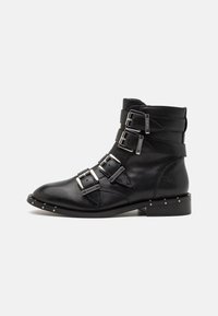 Botas de tobillo de cuero negro con tres correas de hebilla, puntera afilada y acentos de tachuelas plateadas a lo largo de la suela. Tacón plano y textura suave.