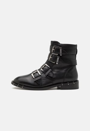 Botas de tobillo de cuero negro con tres correas de hebilla, puntera afilada y acentos de tachuelas plateadas a lo largo de la suela. Tacón plano y textura suave.