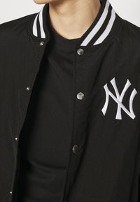 Black varsity jacket with a white striped ribbed collar, metallic snap buttons, and a prominent white embroidered NY logo on the chest.