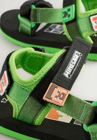 Mustad ja rohelised sandaalid, millel on reguleeritavad rihmad, pehme sisekate ja dekoratiivne Minecrafti tegelane rihmal.