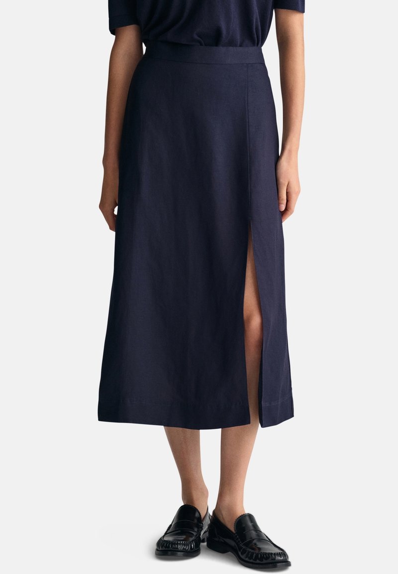 Femme portant une jupe midi bleu marine foncé avec une fente latérale et des chaussures noires sans lacets, debout devant un fond clair uni.