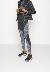 Vrouw die een zwarte donzen jas verstelt, gekleed in zwarte en grijze legging met luipaardprint en zwarte sportschoenen, tegen een witte achtergrond.