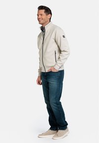Lichtbeige ritsjack met een hoge kraag, twee zijzakken met rits en een zachte textuur, gecombineerd met donkerblauwe jeans en grijze sneakers.