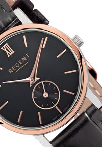 Montre à cadran noir rond avec des accents en or rose, affichant le chiffre romain XII, un petit sous-cadran et un bracelet en cuir noir. Boîtier en métal.