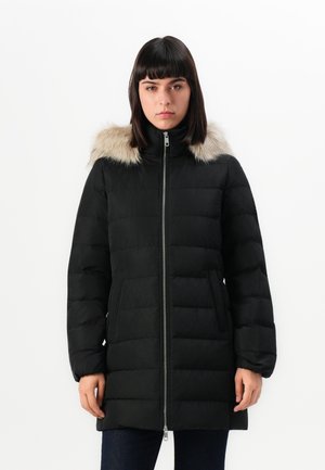 Donna che indossa un cappotto invernale trapuntato nero con cappuccio bordato in pelliccia, in piedi di fronte a uno sfondo bianco uniforme.