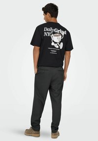 Schwarzes Baumwoll-T-Shirt mit großem weißen Text und Grafik auf dem Rücken. Kombiniert mit dunkelgrauen Hosen und braunen Schuhen.