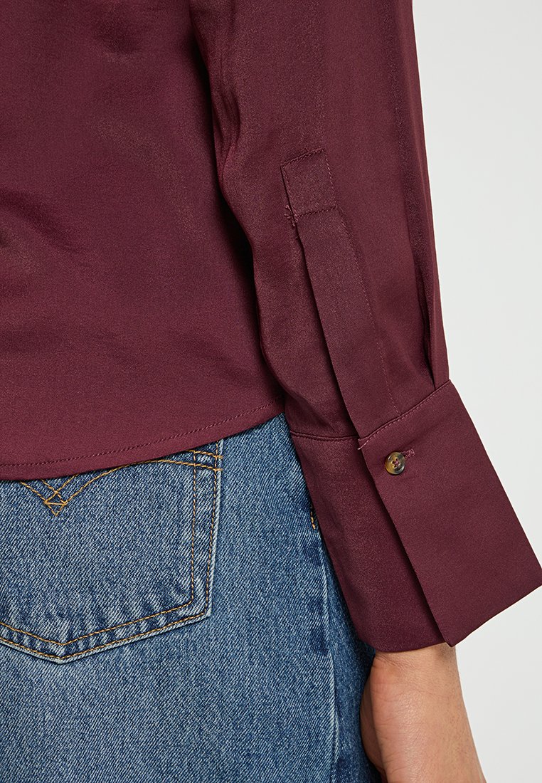 Burgunderfarbige Seidenbluse mit langen Ärmeln, die über Knopfmanschetten verfügt. Kombiniert mit hellblauen Jeans aus Denim, die Details der Gesäßtaschen zeigen.