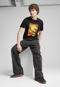 Camiseta gráfica negra con un audaz diseño de gato amarillo sobre un fondo rojo, combinada con pantalones vaqueros oscuros de corte holgado y zapatillas negras.