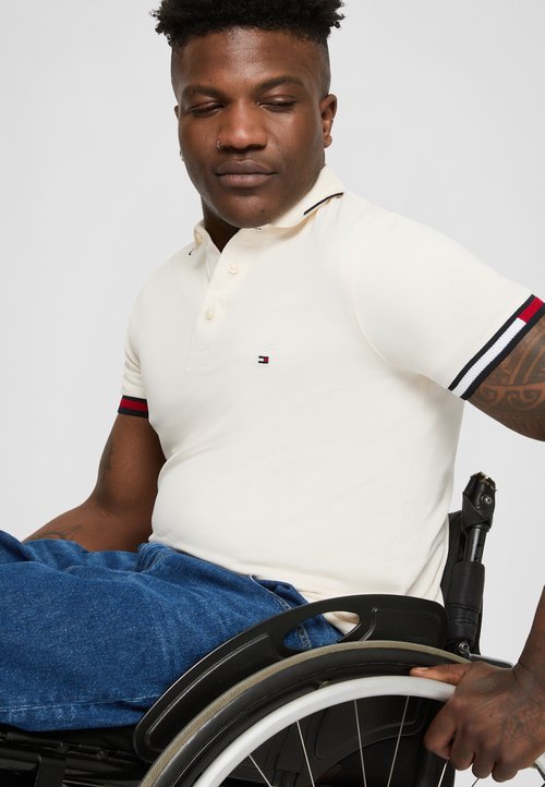 Beige polo tričko s límečkem a třemi knoflíky, s červenými a námořnickými pruhy na rukávech. Nosené s modrými džínovými kalhotami.