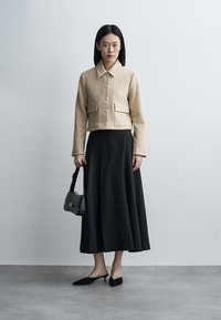 Femme debout contre un mur uni portant une veste beige à carreaux boutonnée, une jupe midi noire, des talons noirs, et tenant un petit sac à main noir.