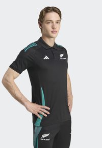 Polo athlétique noir avec des accents en teal sur l'épaule, comportant un logo brodé sur la poitrine et un col structuré.