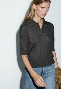 Graues gestricktes Poloshirt mit kurzen Ärmeln, das ein strukturiertes Muster und einen Kragen aufweist, kombiniert mit hellblauen hochgeschnittenen Jeans.